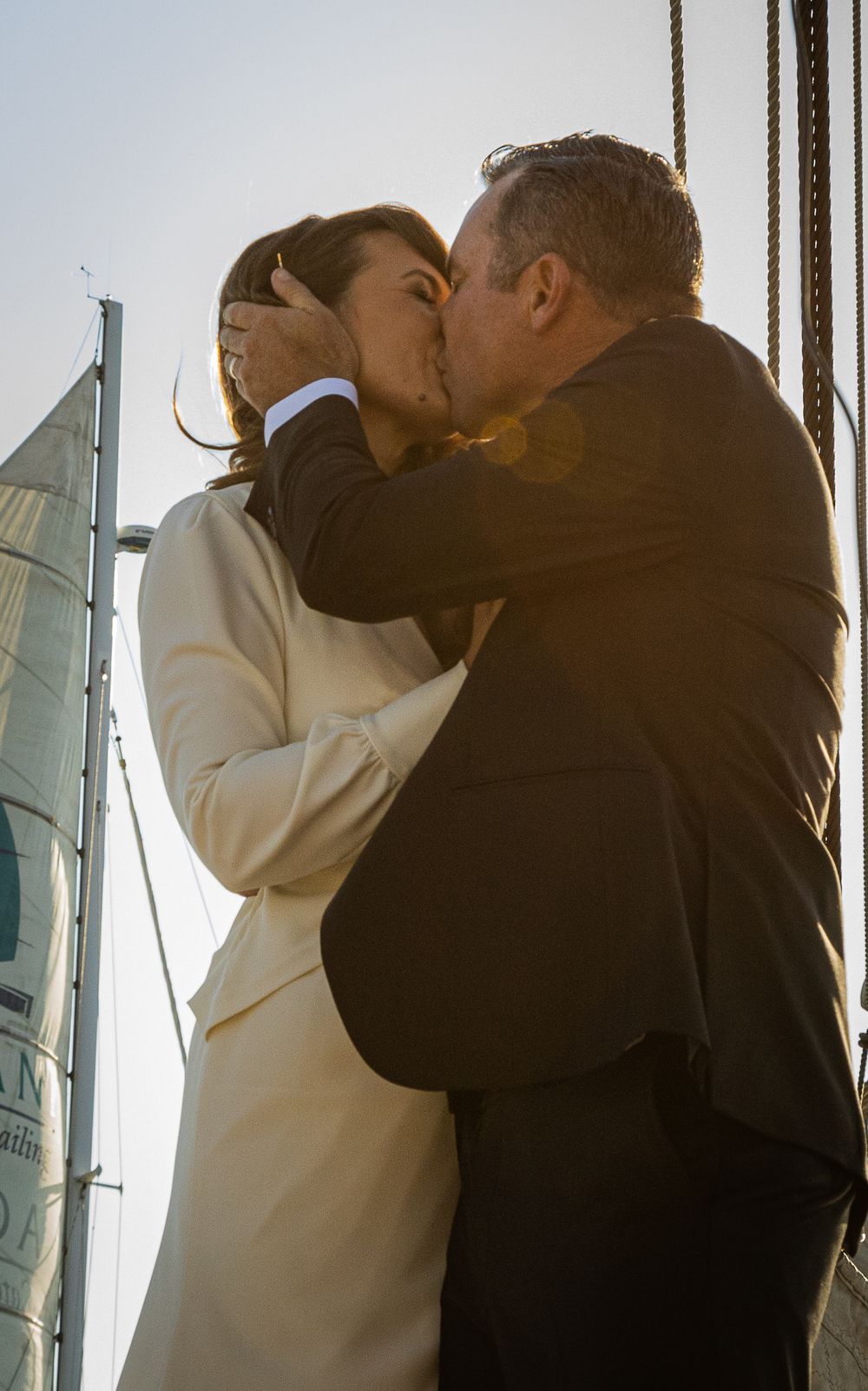 Couple sharing a romantic kiss at sunset on a sailboat, dressed in wedding attire.