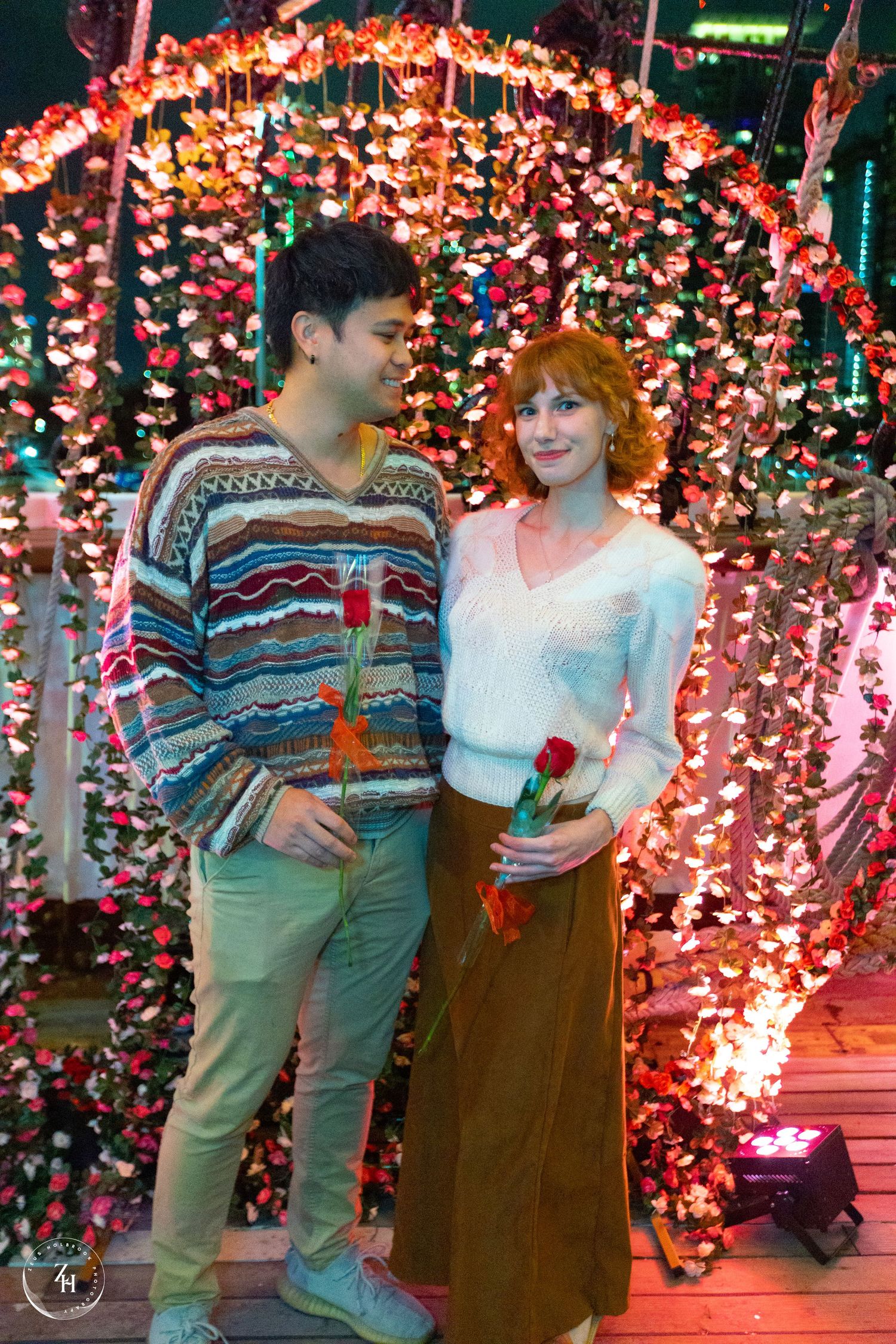 Couple holding red roses standing under a glowing floral arch with colorful lights at a romantic outdoor event.