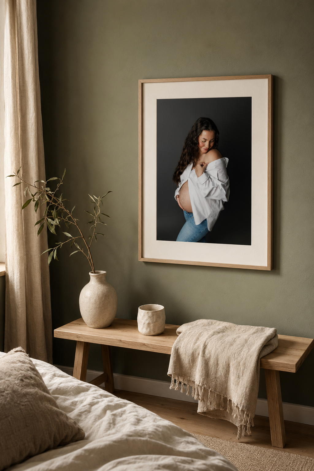 Pregnant woman in white shirt and jeans poses in framed art print displayed in cozy sage green bedroom with wooden bench.