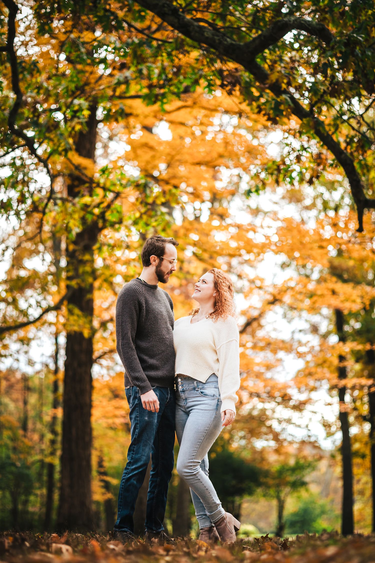Engaged couple standing close together on a wooded trail at Borderland State Park, smiling at each other in golden autumn light.