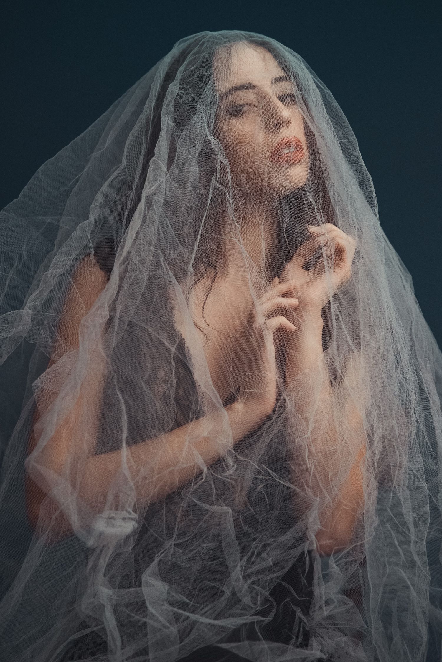 Scatto sensuale e delicato di una donna avvolta in un lungo velo di tulle bianco trasparente, con le mani giunte sul petto e rossetto rosso.