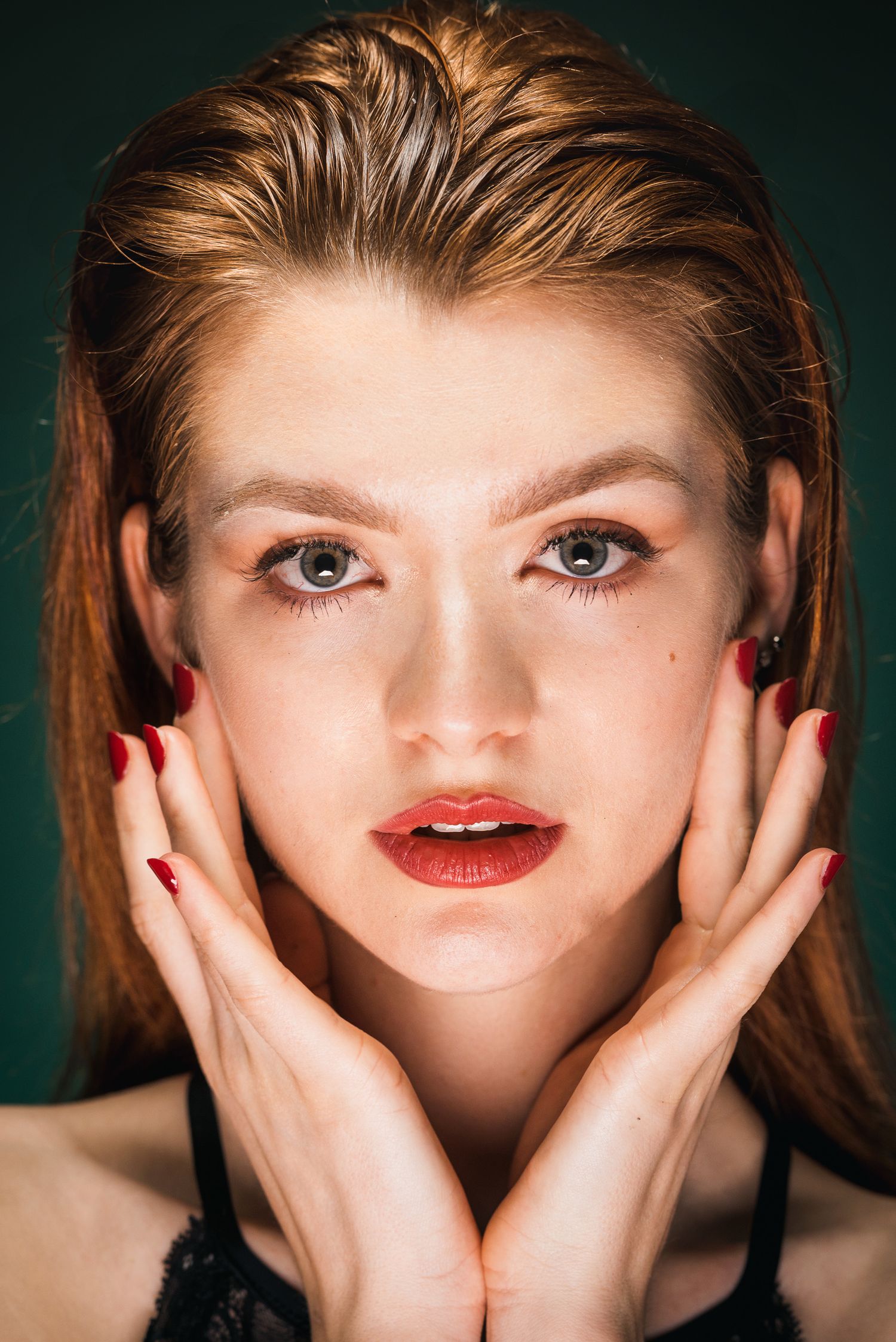 Primo piano ravvicinato di una donna con capelli rossi sfumati all'indietro, rossetto scuro e smalto rosso sulle unghie mentre incornicia il
