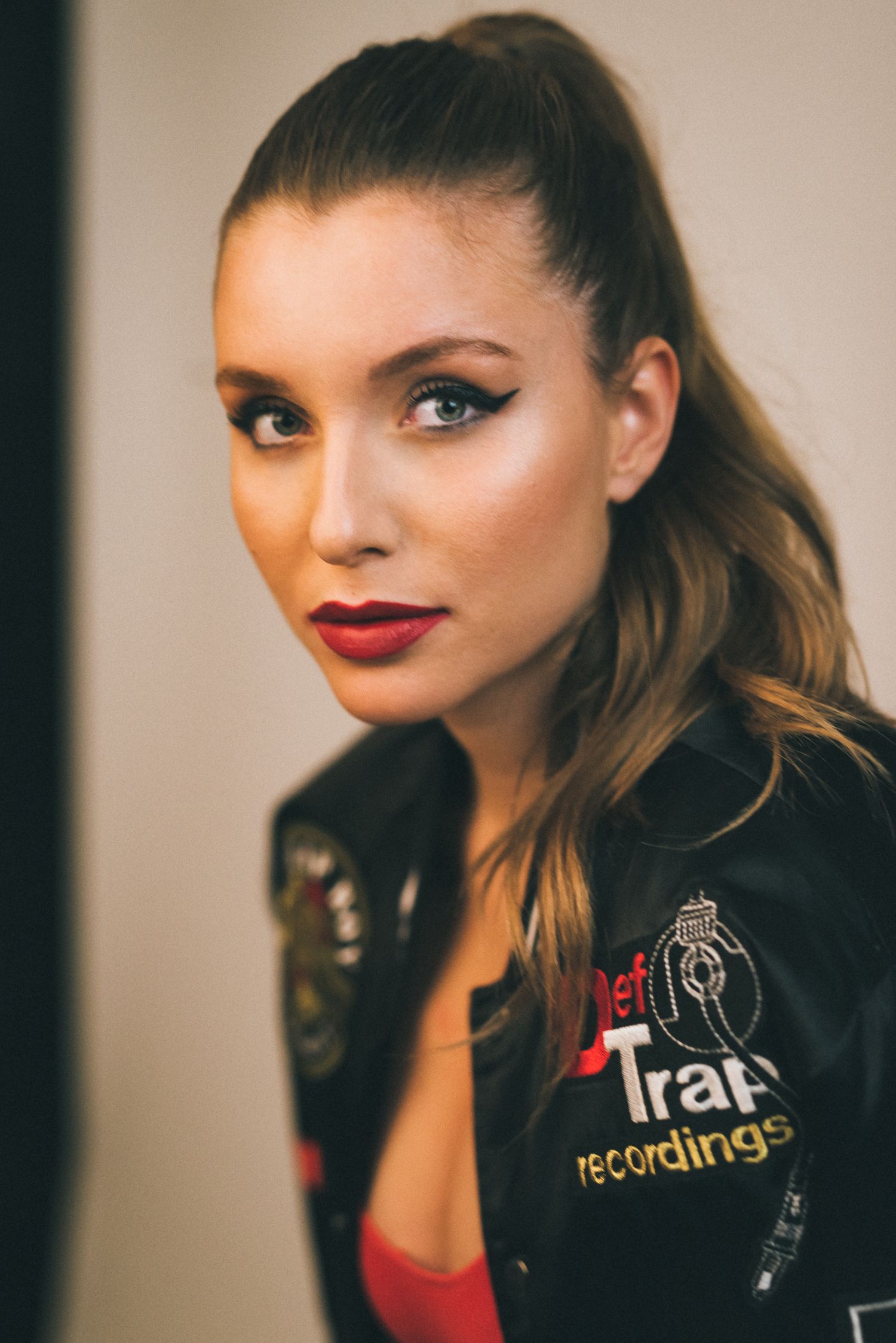 Primo piano di una ragazza con capelli raccolti in una coda alta, eyeliner marcato, rossetto rosso acceso e un giubbotto stile bomber nero c