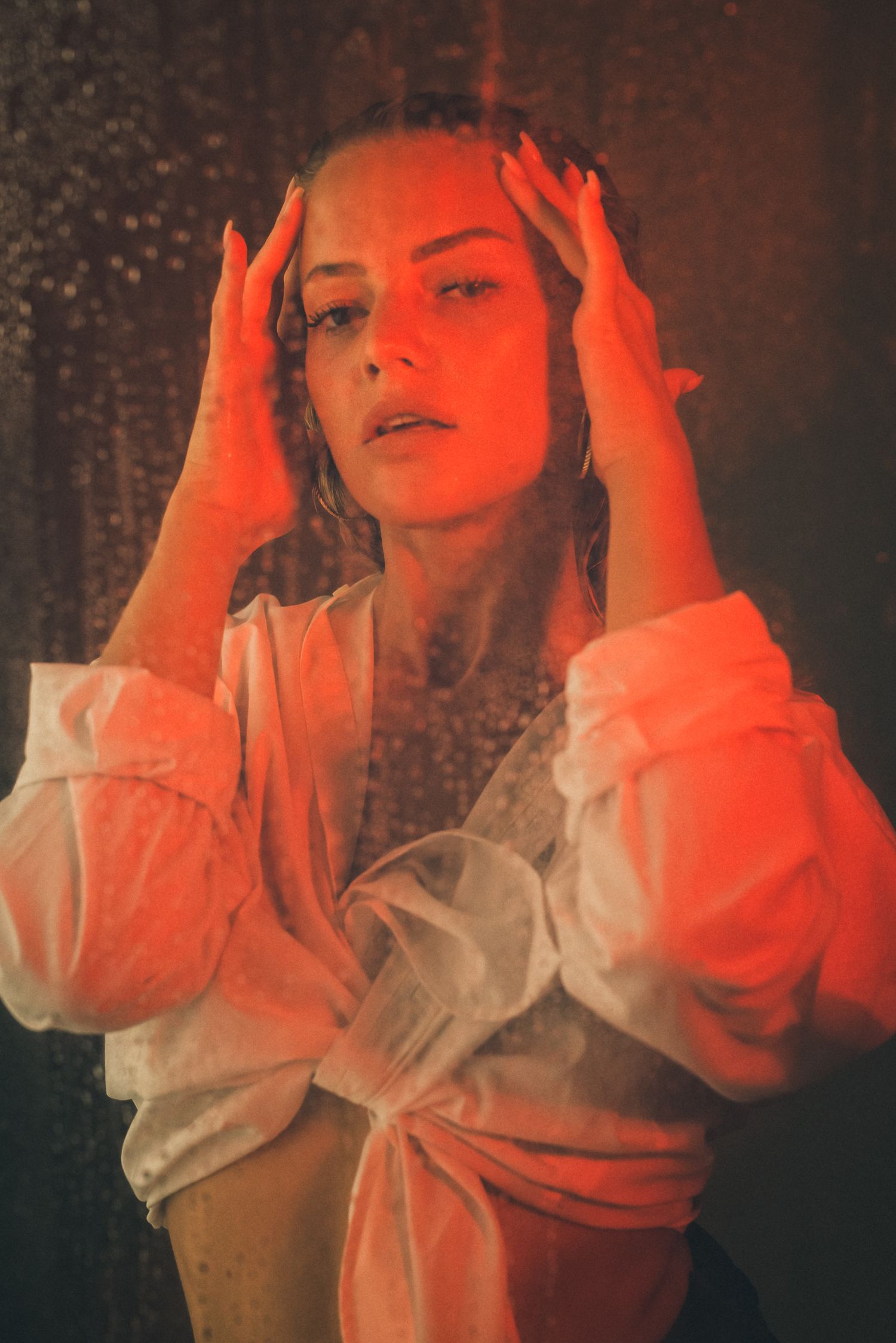 Primo piano espressivo di una donna con camicia bianca annodata in vita, fotografata attraverso un vetro bagnato con un'illuminazione rossa