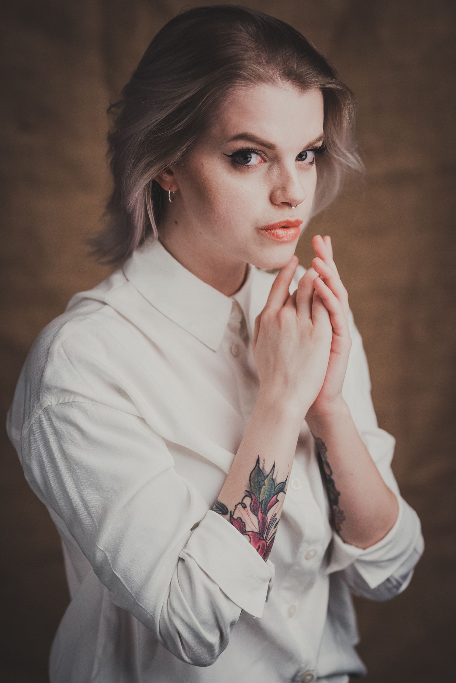 Primo piano espressivo di una donna con capelli biondo platino, camicia bianca elegante e tatuaggio floreale sull'avambraccio, mentre tiene