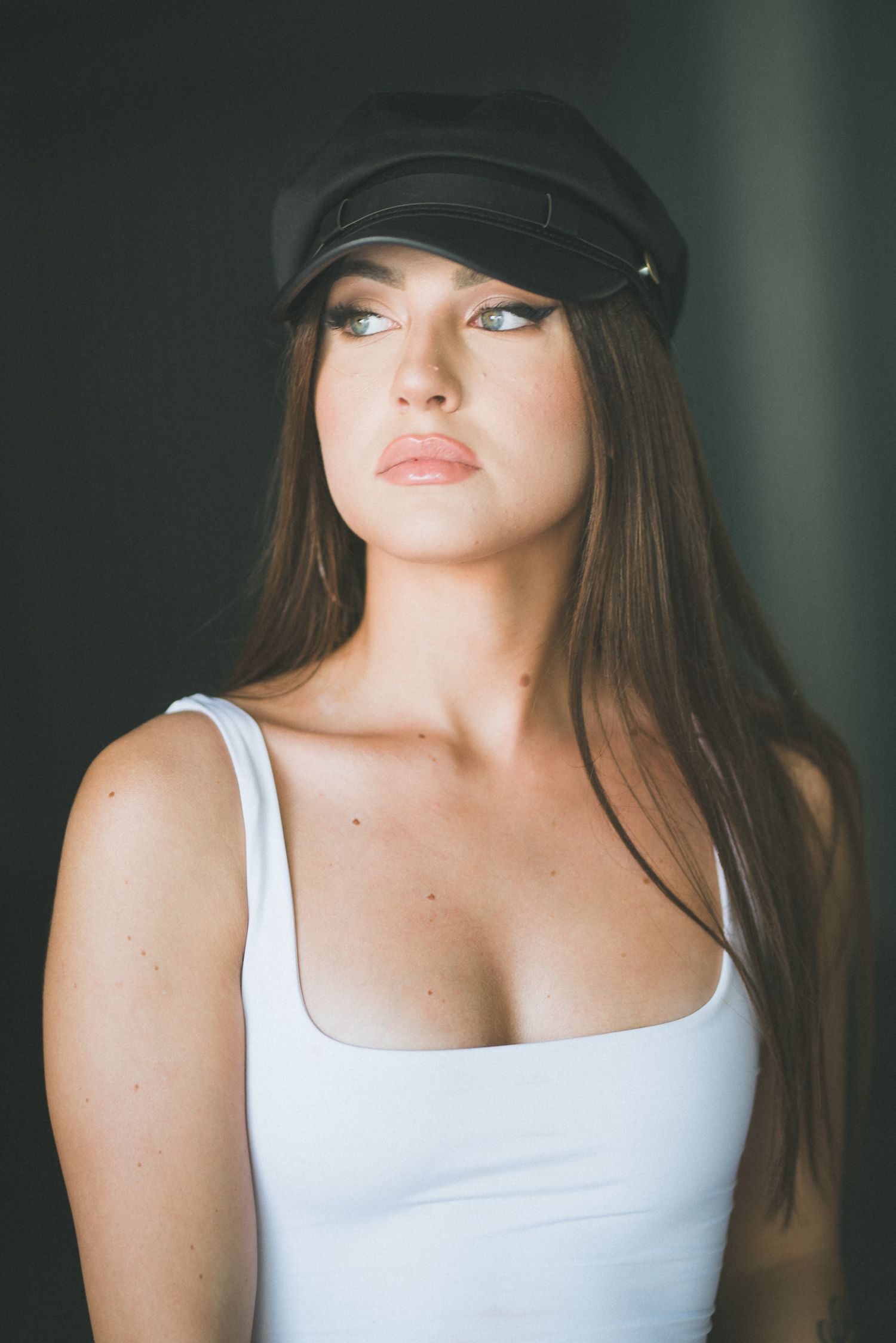 Primo piano di una ragazza con canotta bianca e cappello nero con visiera, mentre guarda leggermente di lato con uno sguardo intenso.