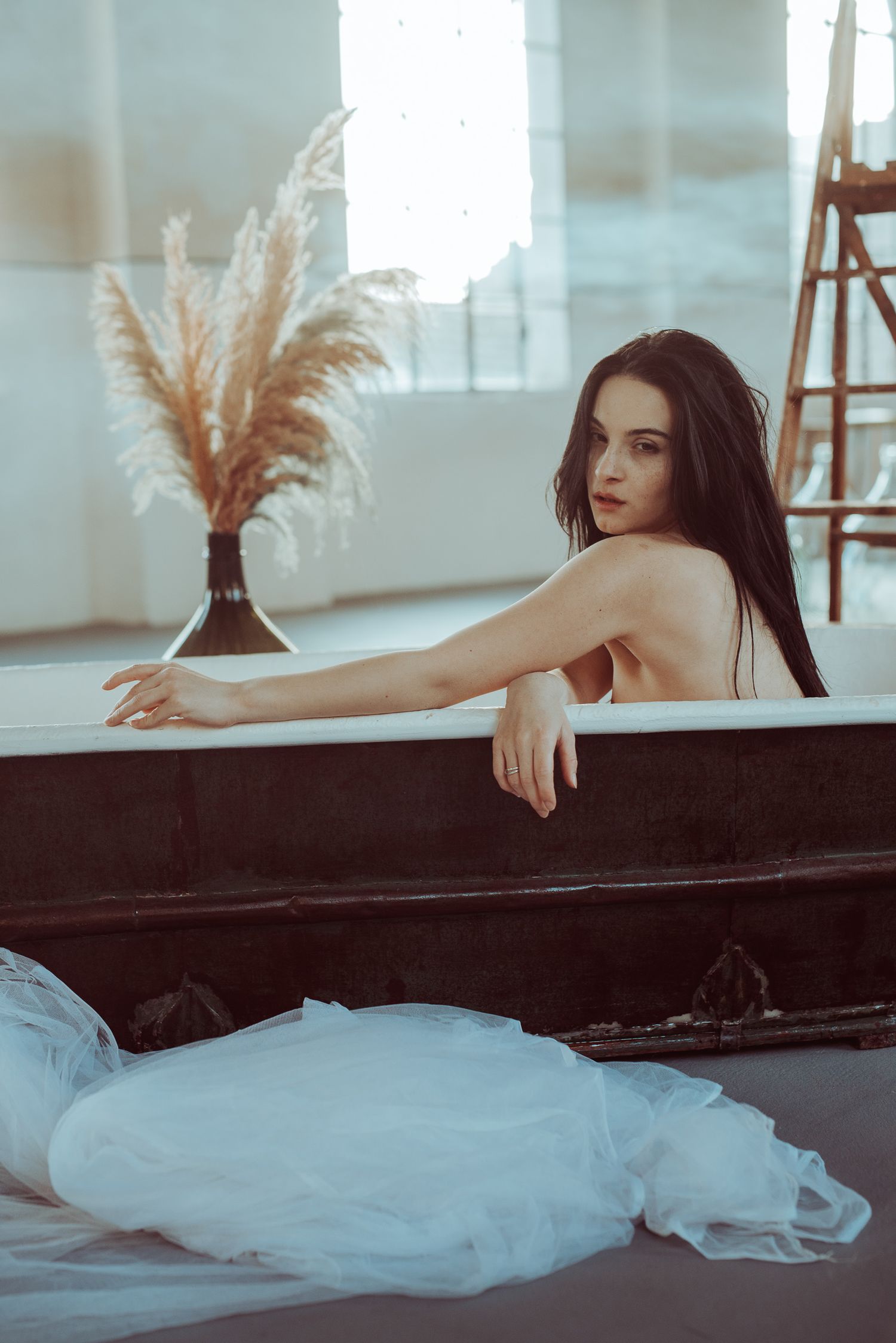 Posa artistica di una donna immersa in una vasca da bagno stile retrò con un velo di tulle bianco adagiato a terra in uno spazio industriale