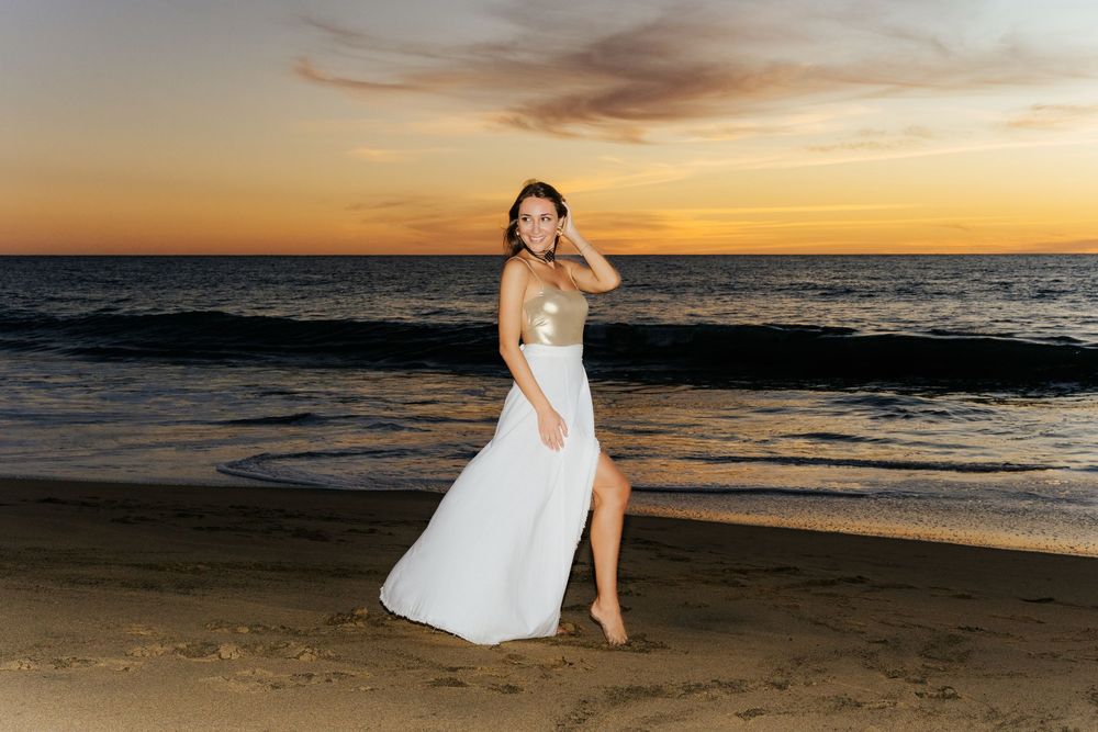 Woman in white and gold gown posing on beach at sunset with ocean waves in background.