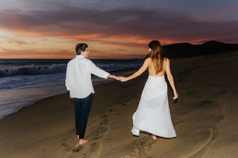 Couple in wedding attire holding hands on a sandy beach at sunset with colorful sky and ocean waves.