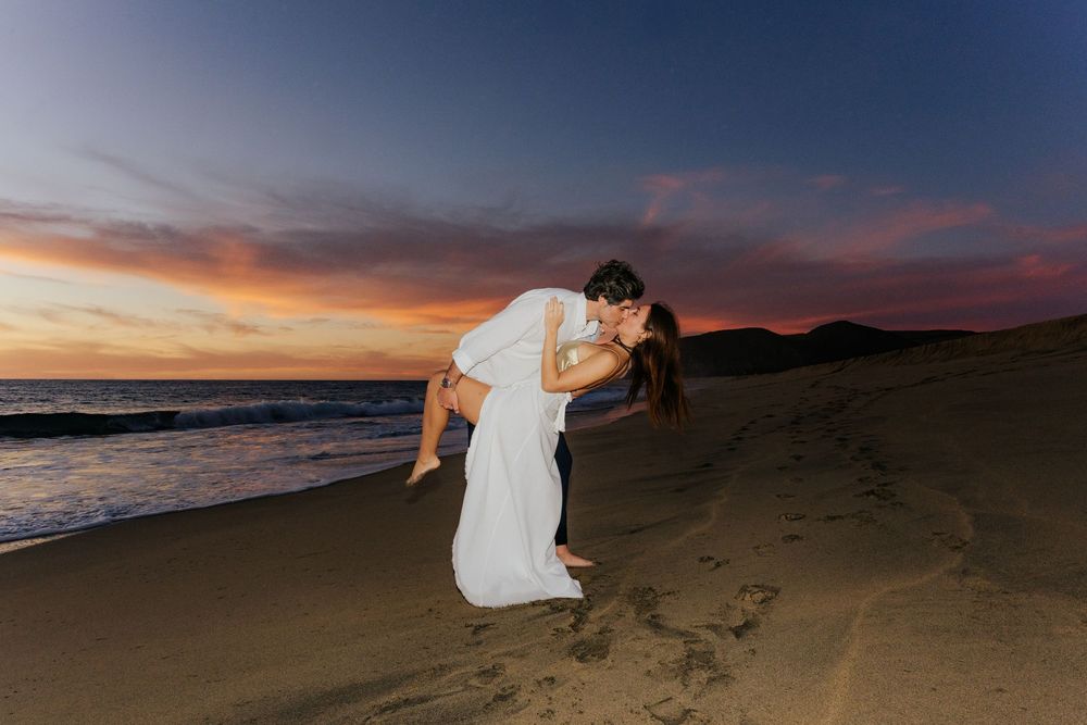 Couple sharing a romantic dip kiss on sandy beach at sunset, waves and colorful sky in background.