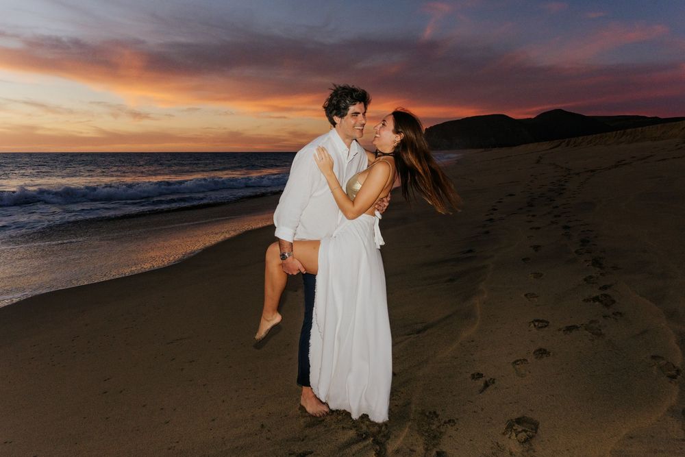 Couple sharing a romantic kiss at sunset on a sandy beach, man lifting woman in white dress.