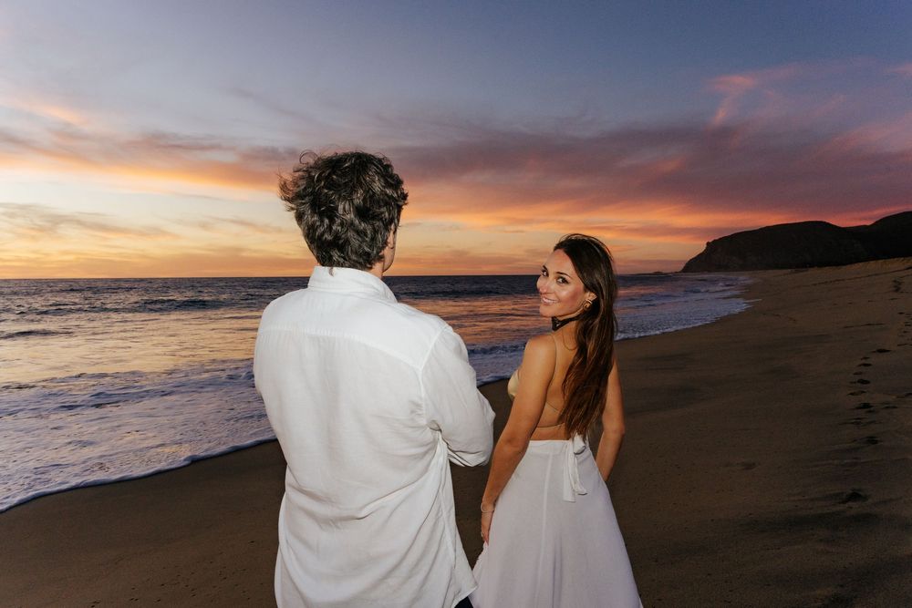 Couple in white attire holding hands on a beach at sunset with vibrant pink and purple sky.