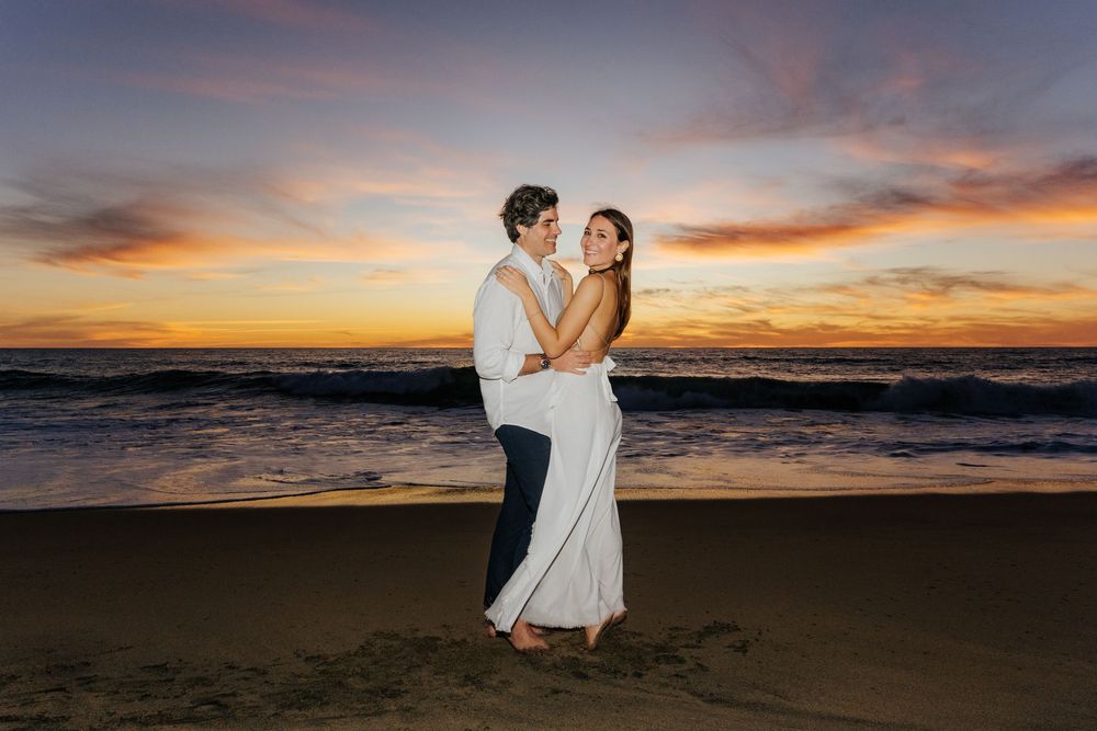 Couple embracing on a sandy beach at sunset with colorful sky and ocean waves in the background.