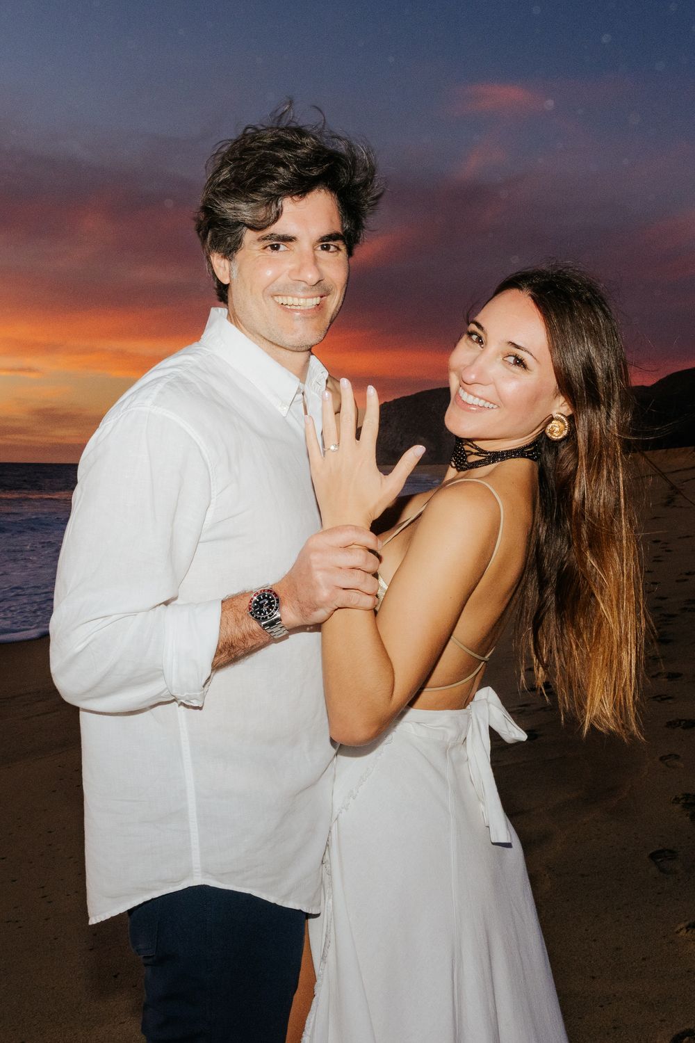 Couple posing on beach at sunset, woman showing engagement ring, both wearing white outfits.