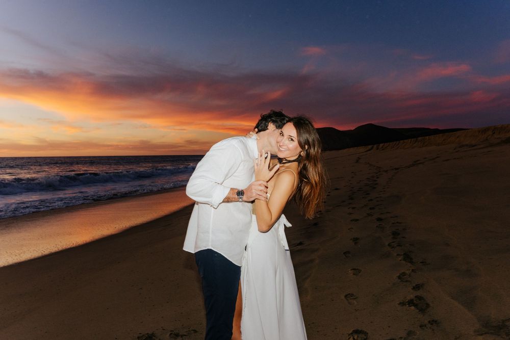 Couple sharing a romantic kiss on a sandy beach at sunset with vibrant pink and purple skies over the ocean.
