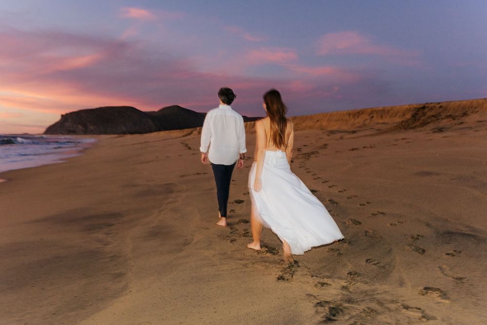 Couple in wedding attire walking hand in hand on a sandy beach at sunset with a colorful pink sky.