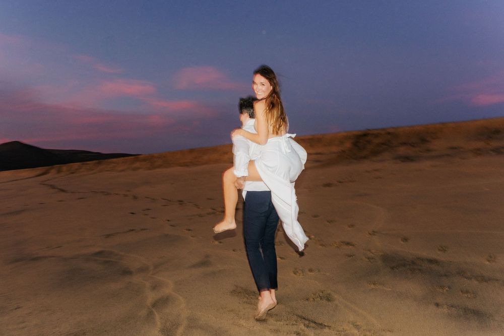 Couple sharing a romantic moment on sand dunes at twilight, man carrying woman in white dress under pink and blue sky.