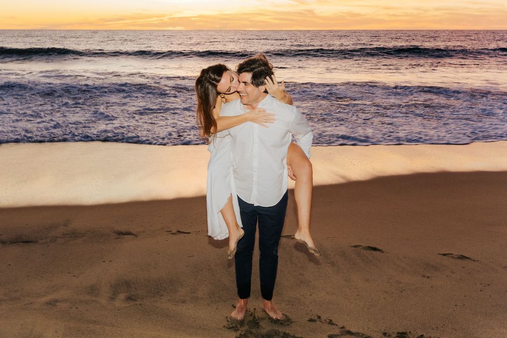 Couple sharing a romantic kiss at sunset on a sandy beach, man carrying woman in white dress.