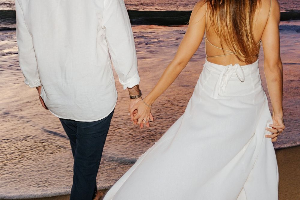 Couple holding hands on beach at sunset, woman in white backless wedding dress, man in white shirt and jeans.