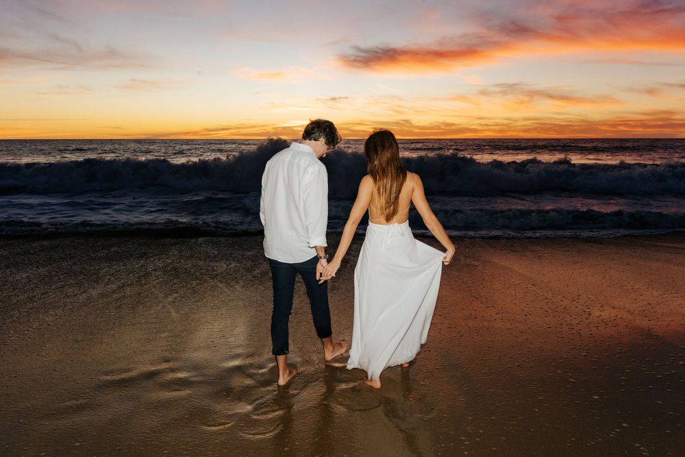 Couple holding hands on beach at sunset, woman in white dress, waves crashing behind them in golden light.