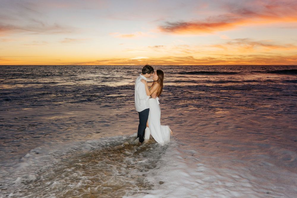 Bride and groom embrace on a wet beach at sunset with vibrant orange and pink skies over the ocean.