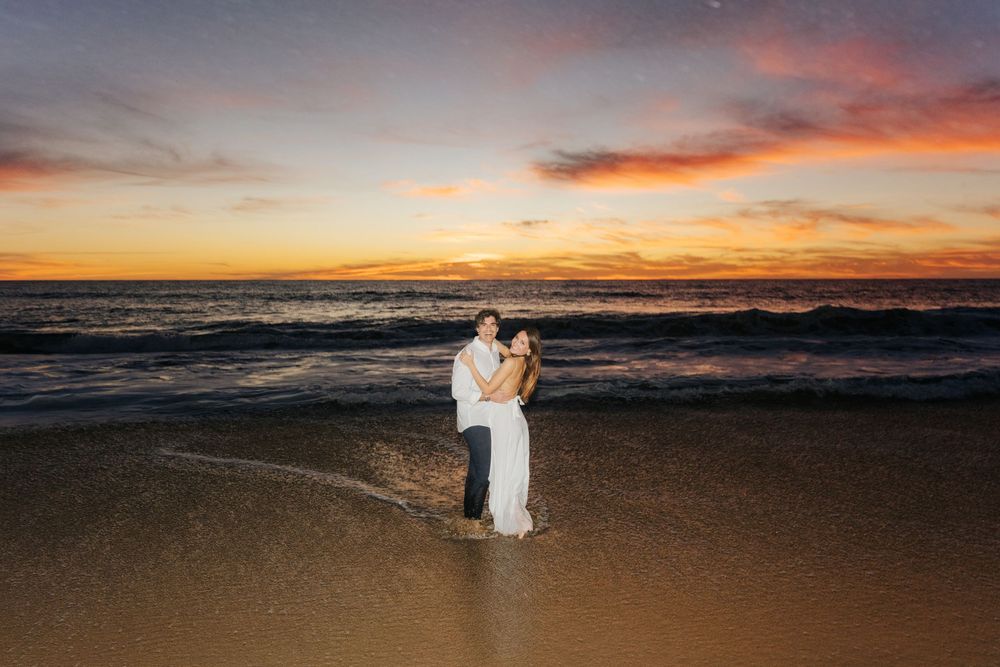 Groom lifting bride on beach at sunset with colorful sky and ocean waves behind them.