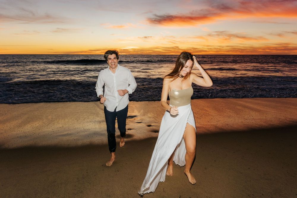 Couple running barefoot on sandy beach at sunset with vibrant orange sky and ocean waves.
