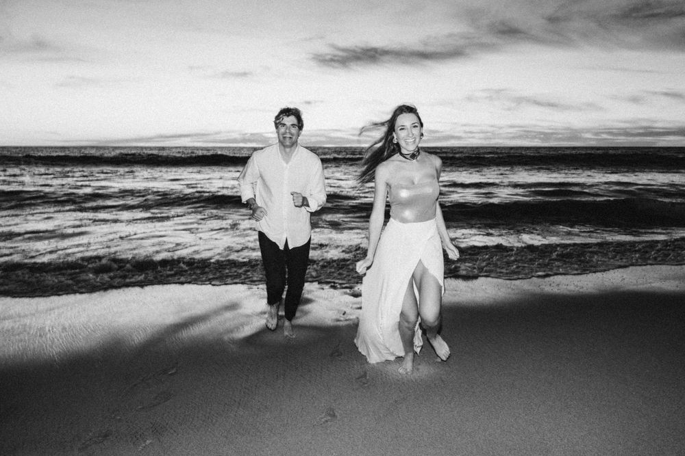 Black and white photo of couple running on beach at sunset with ocean waves behind them.