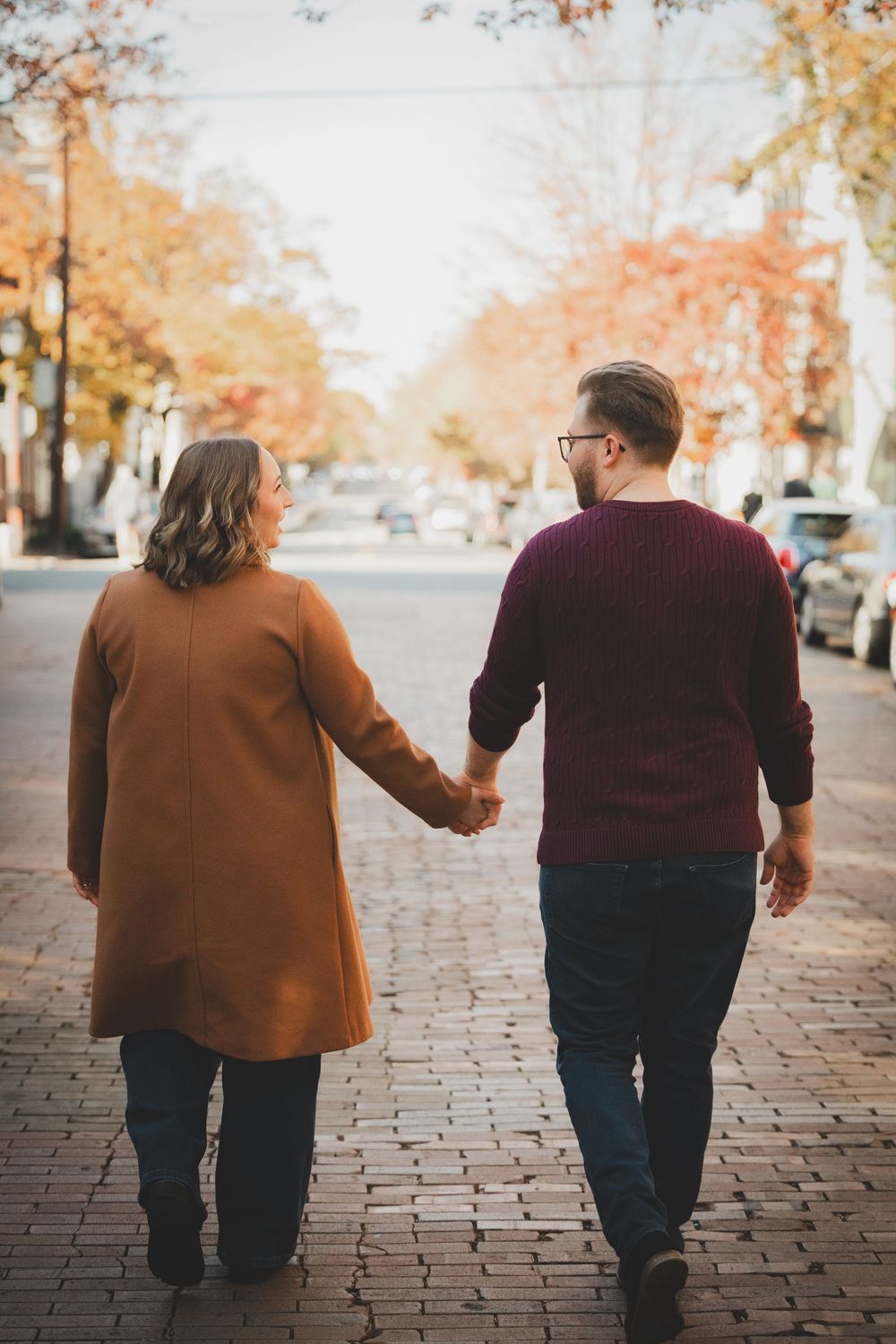Couple holding hands walking down a brick street lined with autumn trees in warm fall colors.