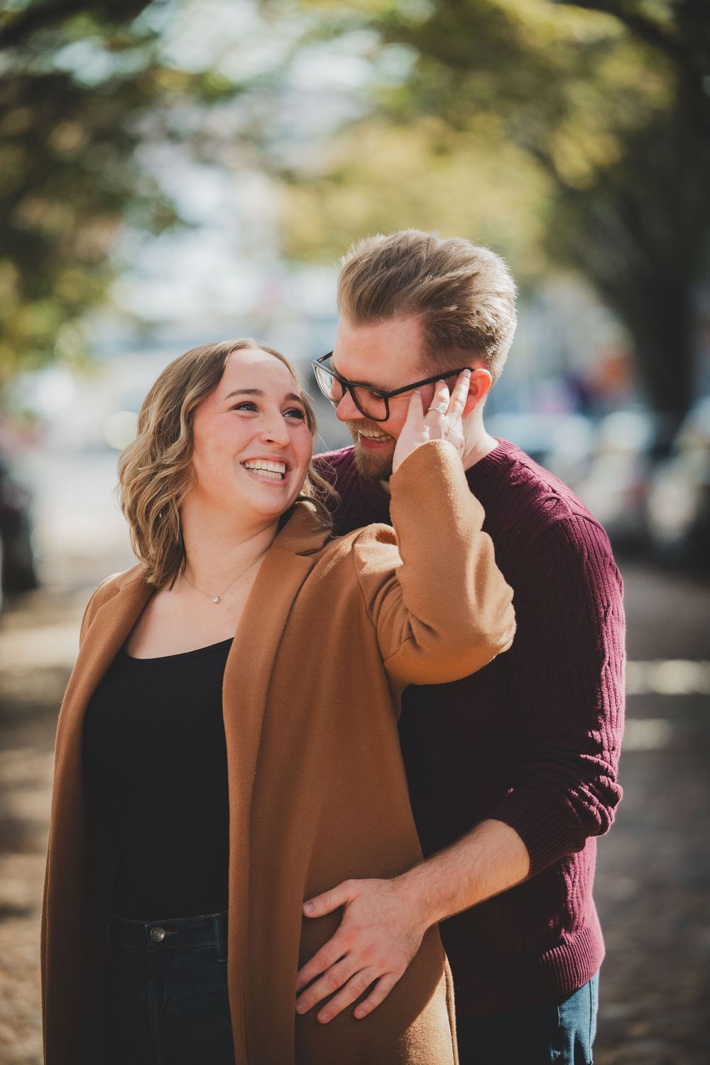 A groom to be embracing bride to be from behind and kissing her cheek while she beams with joy during a fall engagement session