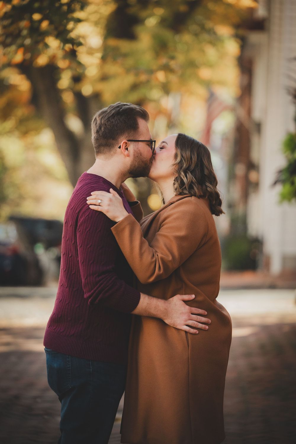 Newly engaged couple sharing their first kiss on a sunlit brick sidewalk with fall trees glowing behind them