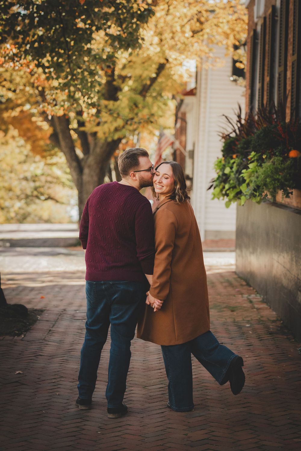 Groom kisses bride's cheek as she laughs joyfully while walking hand in hand on a brick path after their engagement