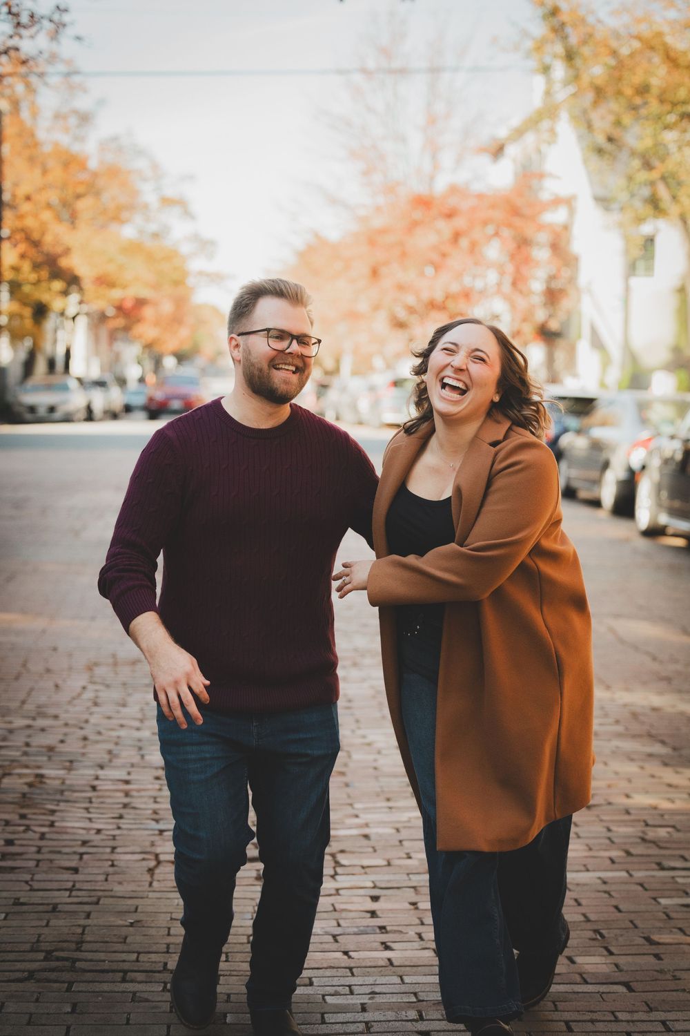 Couple laughing together while strolling down a cobblestone street lined with vibrant autumn foliage in Virginia