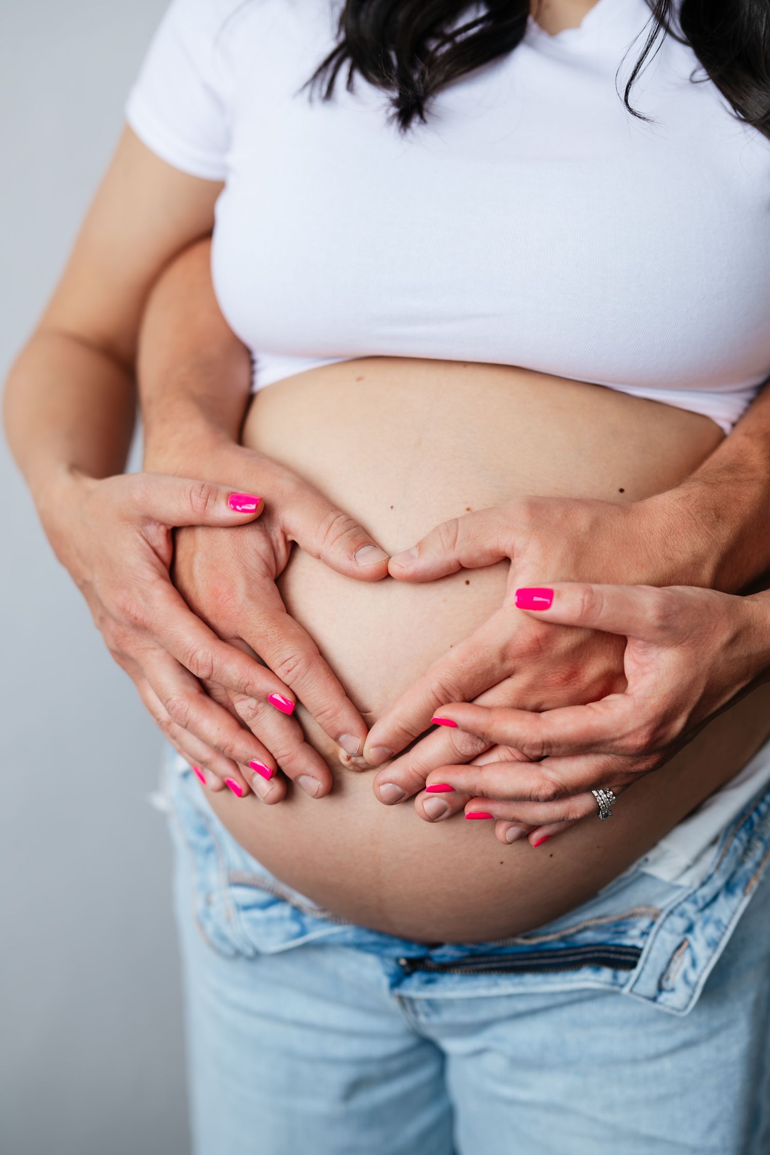 Pregnant woman in white top and jeans with two pairs of hands forming a heart shape on her baby bump.