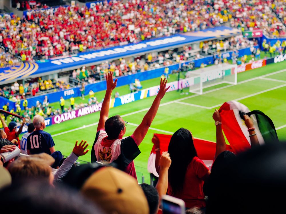 Excited soccer fans cheering with raised hands in a packed stadium during a FIFA World Cup match with Qatar Airways branding.