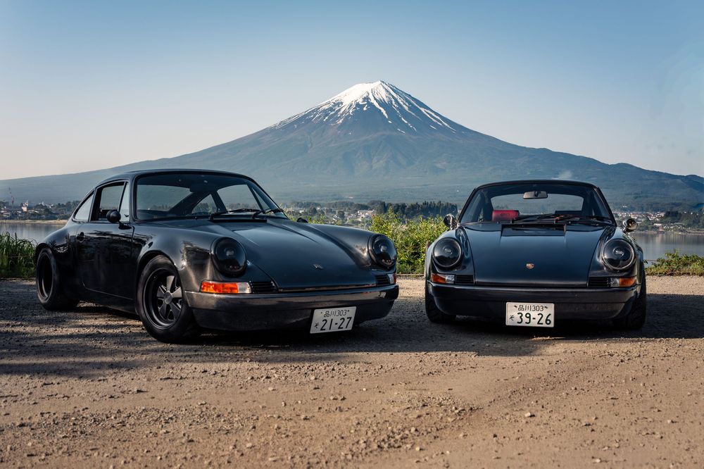 Two classic black Porsche 911s parked with Mount Fuji in the background under a clear blue sky in Japan.