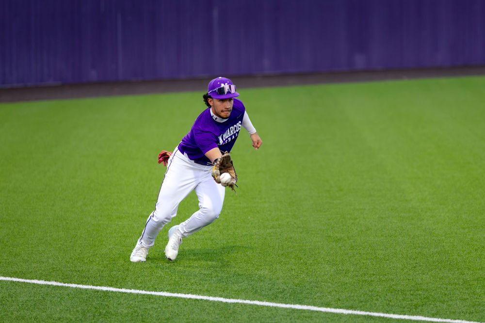 Baseball player in purple uniform fielding a ground ball on a well-manicured grass field during a night game.