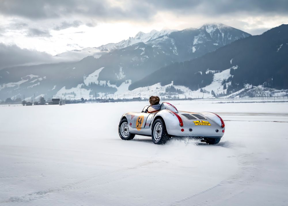 Vintage Porsche 550 Spyder racing on snow-covered alpine track with dramatic mountain backdrop in winter.