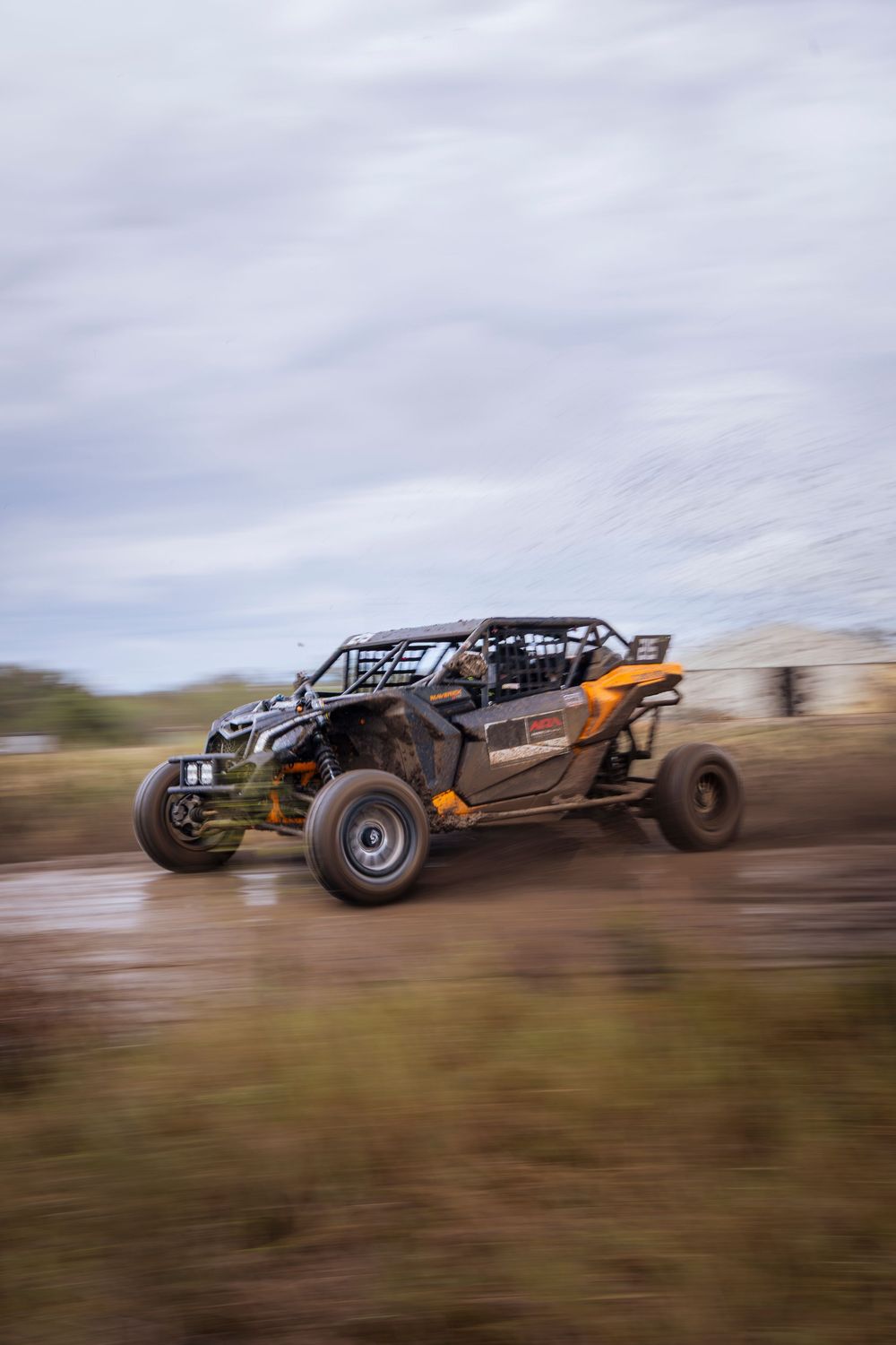 A Can-Am UTV races through muddy off-road terrain at high speed under cloudy skies.