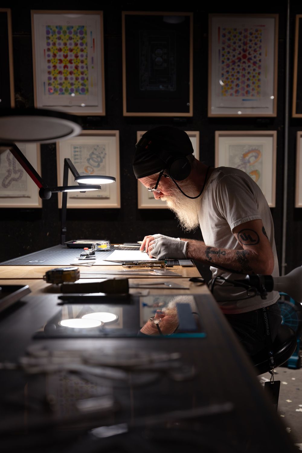 Tattooed artist with beard and beanie works at illuminated desk surrounded by framed artwork in dark studio.