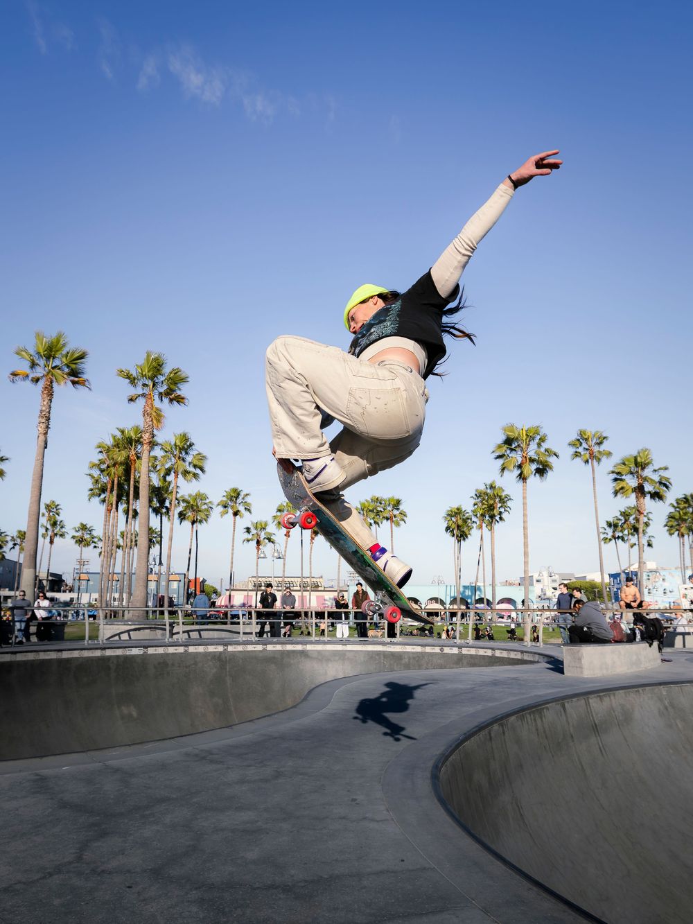 Skateboarder performing an aerial trick at Venice Beach skate park with palm trees and blue sky in background.