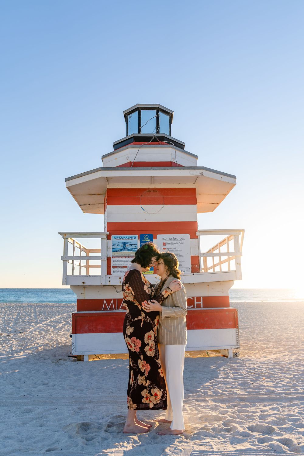 Couple embracing romantically in front of a red and white striped Miami Beach lifeguard tower at sunset.