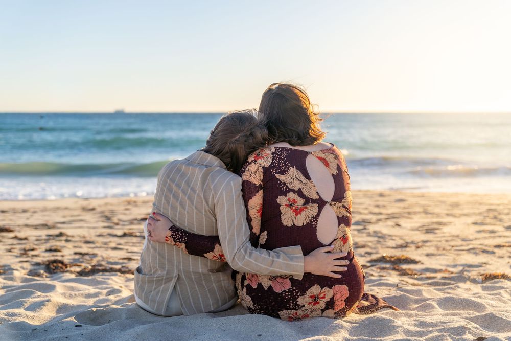 Couple sitting together on sandy beach at sunset, embracing with ocean view in background.