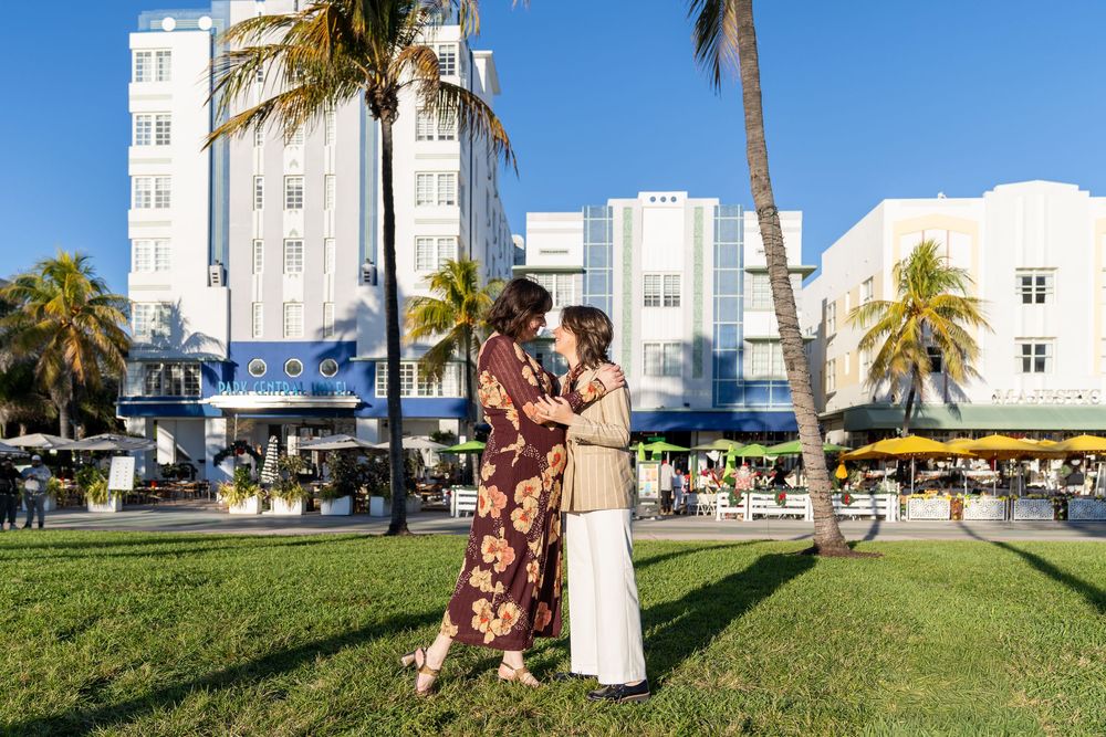 Couple embracing on Miami Beach's Ocean Drive lawn, surrounded by palm trees and Art Deco buildings.