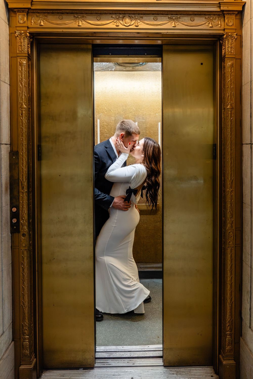 A wedding couple shares a romantic kiss in a vintage elevator with ornate gold trim and brass doors.