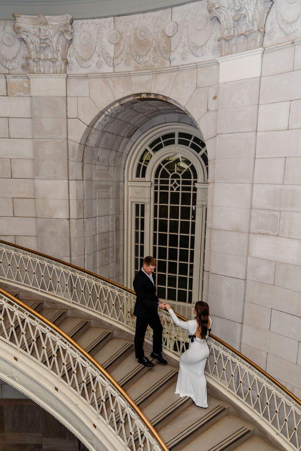 A couple descends an elegant curved marble staircase with ornate railing and arched windows in a grand building.