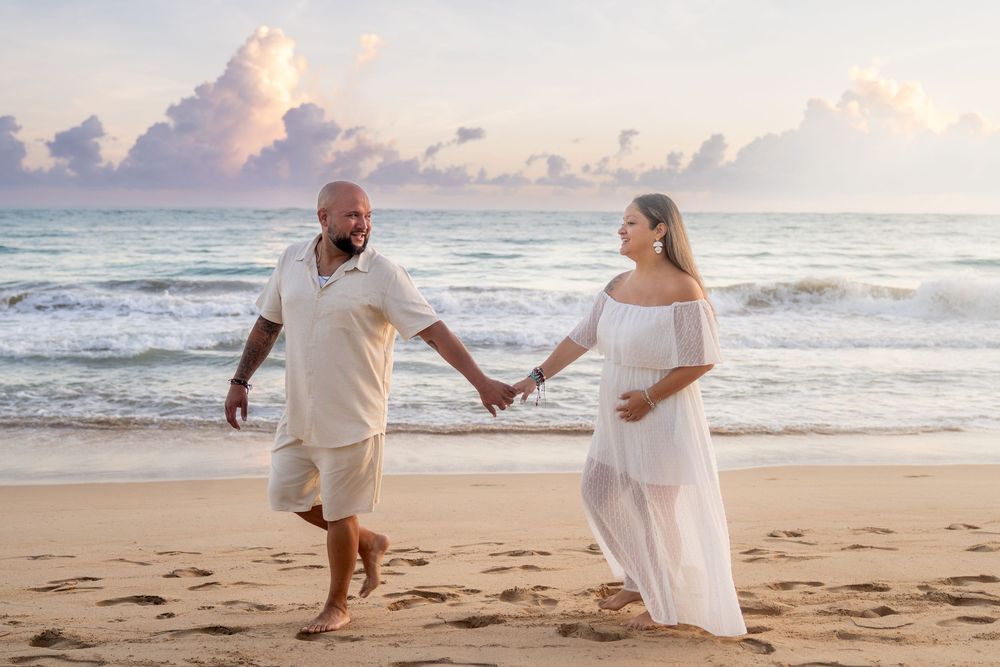Couple holding hands walking on beach at sunset, pregnant woman in white dress, romantic maternity photo.