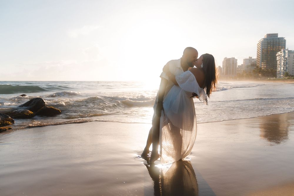 Couple sharing a romantic kiss on a sunlit beach with ocean waves and city skyline in the background.
