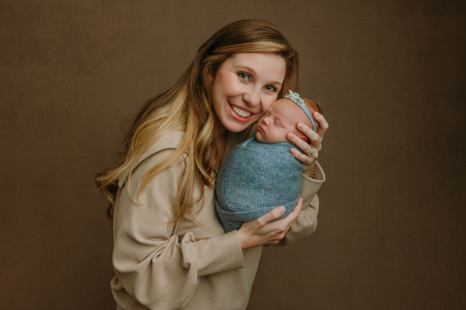 A mother hugs a newborn baby girl while smiling warmly in a dimly lit setting at the newborn photography studio in Victoria BC.