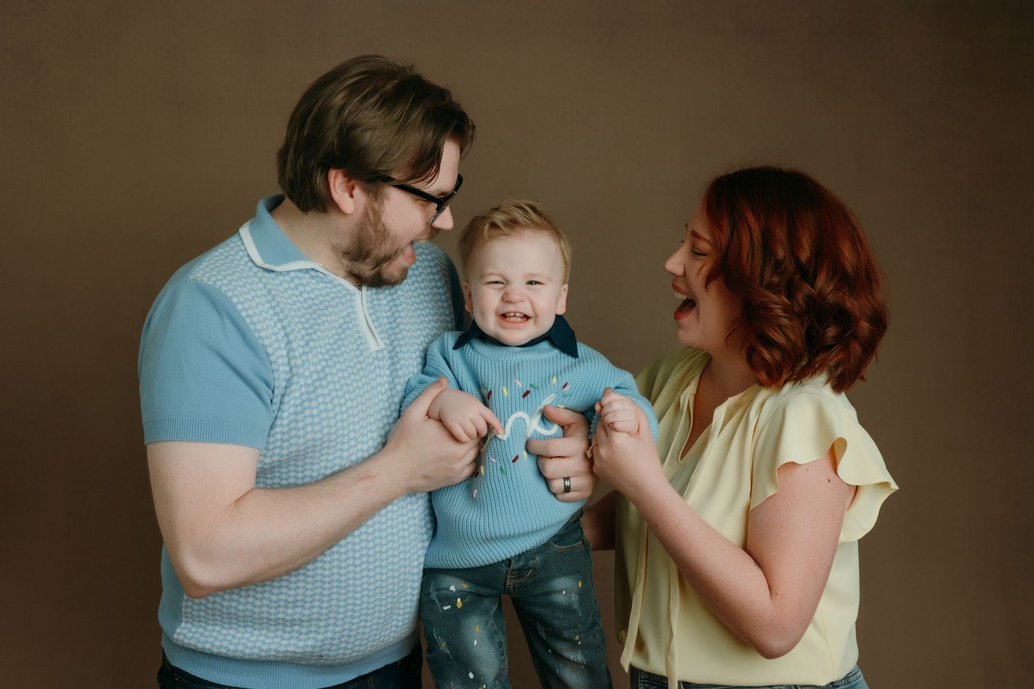 Happy family portrait with parents and toddler boy in blue sweater laughing together at the family portrait specializing studio in Victoria